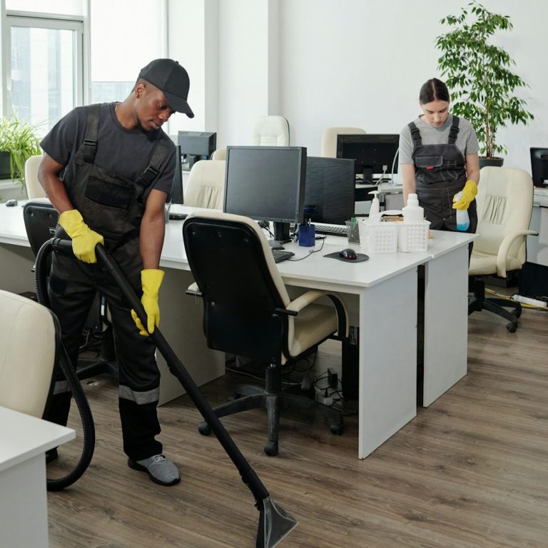 Zwei Reinigungskräfte säubern ein modernes Büro mit Staubsauger und Reinigungsmitteln.